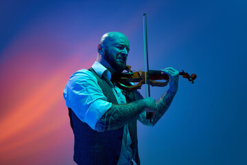 Deep feelings of melody. Young bald man playing violin against blue background in neon with mixed light. Concept of music, talent show, performance, concert, festival, instruments