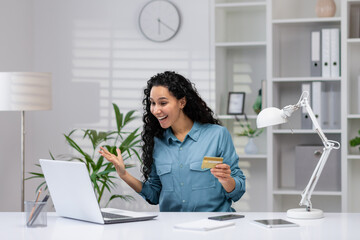 Joyful Hispanic woman with credit card making purchases online while working from home office, expressing excitement.