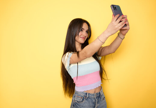 Happy Beautiful young brunette woman using smartphone mobile to selfie take photo herself standing on isolated yellow background.