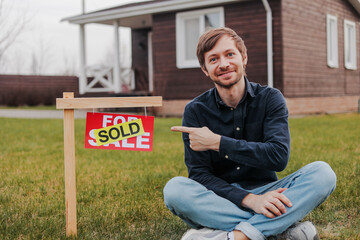 Portrait of smiling young man pointing to sold sign outside house. Happy caucasian man buying new home. - obrazy, fototapety, plakaty