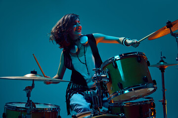 Beautiful, energetic young woman, artistic musician playing drums against cyan background in neon light. Concept of music, talent show, performance, concert, festival, instruments