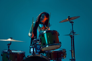 Artistic, expressive young woman, musician playing drums against cyan background in neon light. Concept of music, talent show, performance, concert, festival, instruments