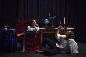 Creative portrait of two medieval persons sitting in room with vintage furniture and drinking wine while writing novel. Concept of comparisons of eras, renaissance, friendship, inspiration. - obrazy, fototapety, plakaty