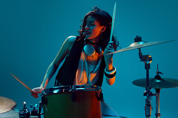 Beautiful young woman in stylish clothes, musician playing drums against cyan background in neon...
