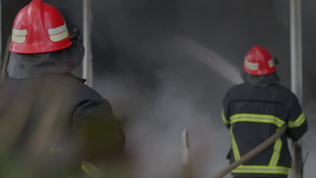 A fireman in helmet and safety uniform extinguishing the fire with hose while another one watches him in the background. Concept of home insurance, public safety, 911 emergency, dangerous work