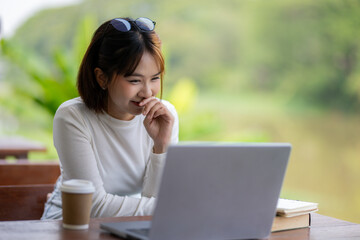 A smiling woman engaging with a laptop at an outdoor table by the riverside, with a coffee cup and...
