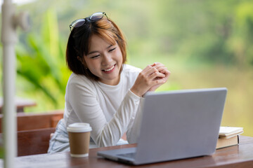 A smiling woman engaging with a laptop at an outdoor table by the riverside, with a coffee cup and...