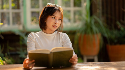 A young woman deeply absorbed in reading a book, bathed in warm sunlight at a rustic outdoor table,...