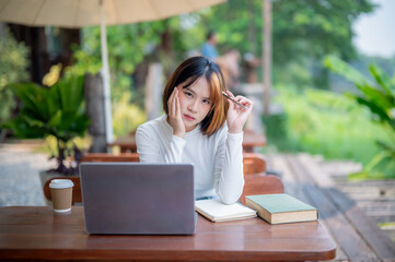 Young woman wearing glasses sitting at a garden table, looking thoughtful with a laptop and notebooks in front of her.
