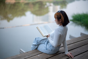 A casual young woman smiling at the camera while enjoying a book on a wooden pier by the water,...