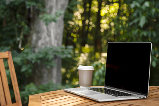 A paper cup of coffee and a laptop on a wooden table on a blurred forest background, work in nature, on vacation and remotely, copy space.