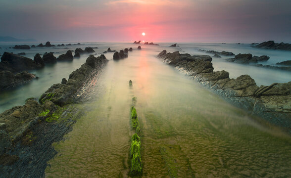 A fiery sunset casts a warm glow over the moss-covered rocks and silken waters of Playa de Barrika in Bilbao, creating a surreal seascape