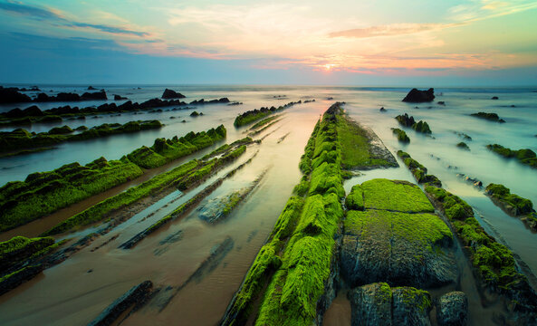 The sun dips below the horizon, casting a soft glow over the moss-covered rocks at Playa de Barrika, Bilbao, against a silky sea