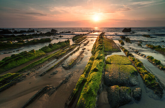 The setting sun bathes Playa de Barrika in golden light, highlighting the textured mossy rocks that lead into the sun-kissed sea