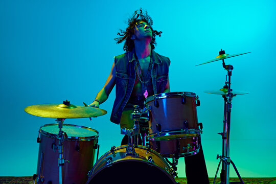 Stylish young man, musician playing drums, making solo performance against blue background in neon light. Concept of music, talent show, performance, concert, festival, instruments, youth culture