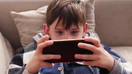 Caucasian boy 9 -10 years old lying on the sofa at home uses a smartphone, playing mobile games, watching his favorite content. teenager plays mobile games at home