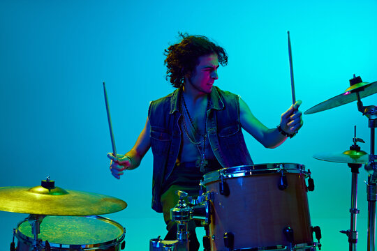 Enthusiastic young boy, drummer with curly hair playing drums against blue background in neon light. Concept of music, talent show, performance, concert, festival, instruments, youth culture