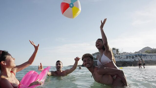 A group of cheerful young friends in swimwear enjoying leisure time on the shore on the sea, playing with a beach ball under the blue sky. Gen z people having fun in summer vacations