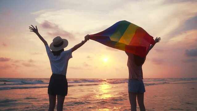 Happy lesbian couple waving lgbt flag on sunset light in beautiful beach. Happy asian lesbian woman demonstrate their rights. LGBTQI, Pride Event, LGBT Pride Month, Gay Pride Symbol