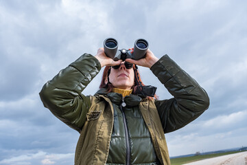 female ornithologist birdman watches birds with binoculars on dirt gravel road against a background...