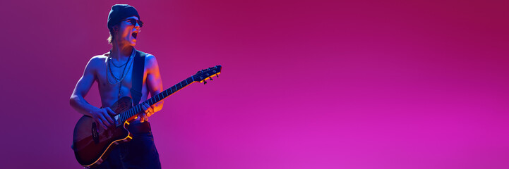 Emotional shirtless young man in sunglasses and hat playing electric guitar against pink background...