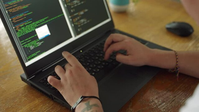 Tattooed hands type on laptop keyboard, coding software. Developer programs app, debugs code, tech workspace scenario. Creative coder at work, programming, developing applications. Slow motion.