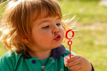 A pretty two-year-old blonde girl plays with blowing soap bubbles on a sunny day
