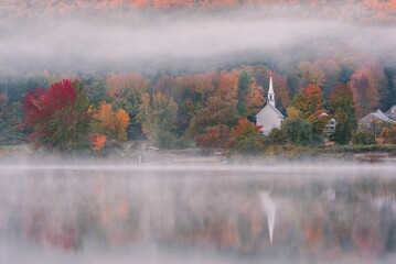 Beautiful view from a lake by autumn trees and a church in misty weather