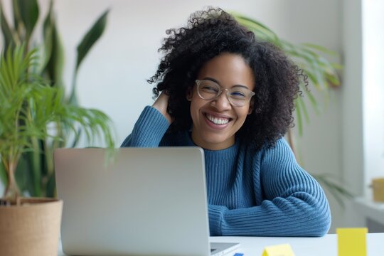 An Attractive Black Woman Of 30 Years Old, Wearing A Blue Sweater And Glasses, Smiling And Looking Happy, Sitting At A Table With A Laptop Surrounded By Plants, And Working In A Home Office