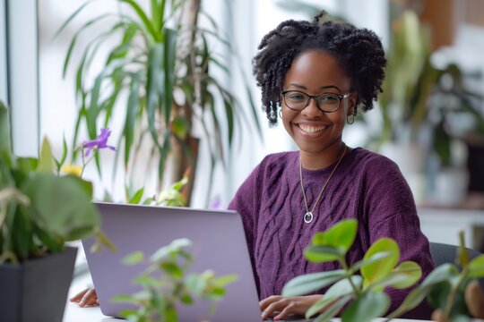 An attractive black woman of 30 years old, wearing a purple sweater and glasses, smiling and looking happy, sitting at a table with a laptop surrounded by plants, and working in a home office