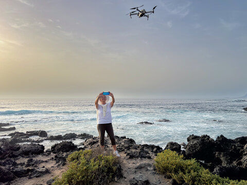 Active elderly woman on a rocky beach takes photos of the drone flying above her. Horizon over the sea