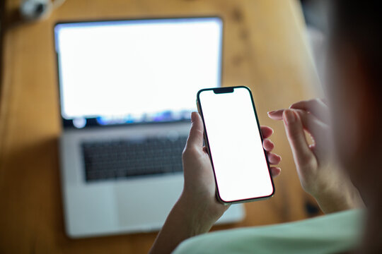 Woman holding smartphone with blank screen in focus laptop in background