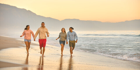 Couple With Friends On Vacation Holding Hands Running Along Beach Shoreline At Dawn