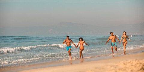 Couple With Friends In Swimwear On Vacation Holding Hands Running Along Beach Shoreline 