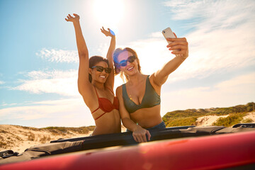 Two Women On Vacation In Car On Road Trip To Beach Standing Through Sun Roof Posing For Selfie