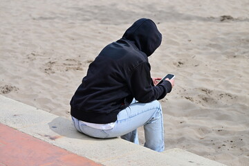 A young girl hidden under her black hoodie while watching her smartphone on a warm, but stormy day at the beach of Benidorm-Spain. 