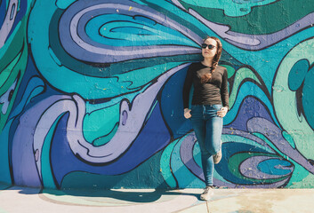Relaxed young female in jeans and sunglesses near multicolored graffiti wall on Belgrade city street, Serbia
