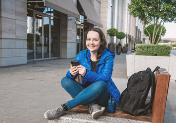 Beautiful smiling tourist woman with backpack sitting on the bench with mobile phone in the city