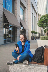 Beautiful young smiling tourist woman with backpack sitting on the bench with mobile phone in the city