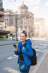 Beautiful smiling tourist woman with mobile phone and backpack standing near road in the Belgrade city, Serbia
