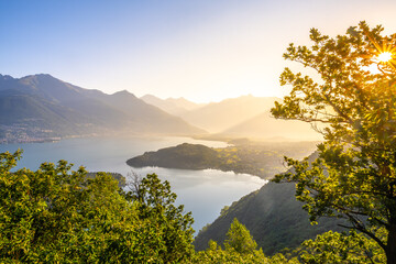 A beautiful mountain range with a lake in the foreground. The lake is calm and the mountains are covered in trees. The sky is clear and the sun is shining brightly. The scene is peaceful and serene - 774146271