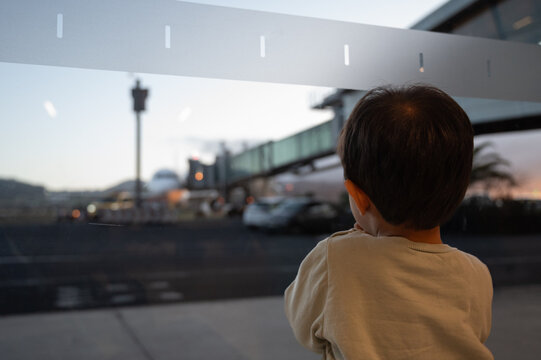 A young child is looking out a window at a busy airport. The scene is bustling with activity, with planes taking off and landing, cars driving on the road, and people walking around