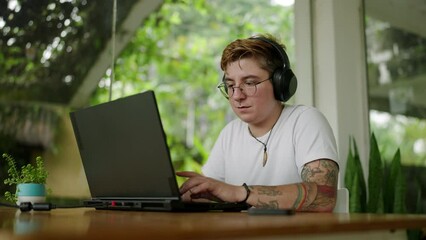 Transgender editor with headphones works on video project in coworking space. Inked professional edits film on laptop, comfortable open office environment. Inclusive workplace, adaptive tech use.