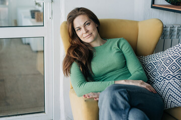 A gorgeous long-haired red-haired woman sitting in a chair calmly looks at the camera.