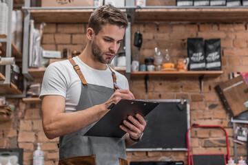 Young owner in an apron writing on clipboard while checking products in restaurant. Man looking on checklist and doing inventory. Small business concept. Entrepreneur in hospitality industry.