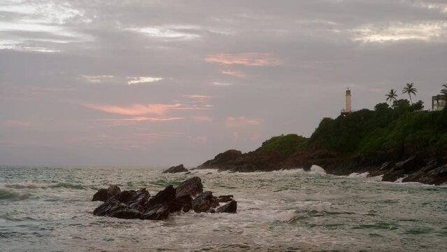 Waves crash against rocks on tropical coastline at dusk. Ocean spray, rise and fall on rough sea. Cloudy sunset sky colors over palm-fringed shore, lighthouse backdrop. Coastal nature scene, tranquil.