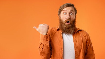 Surprised redhaired man with beard pointing thumb finger aside, studio