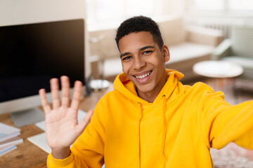 Cheerful black teen waving taking selfie while studying at home