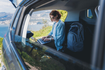 Young woman sitting in the open trunk of a car overlooking the sea and using a laptop, freelancer, remote work and summer vacation auto travel - obrazy, fototapety, plakaty