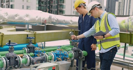 Two engineers with hard hats are checking the gauge on a pipeline valve in an urban industrial